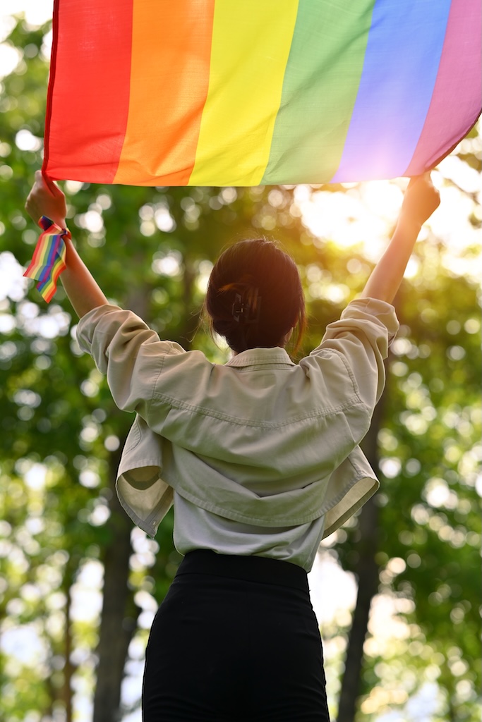 Rear view of young woman holding and raising a rainbow flag with sunlight shining through green trees.