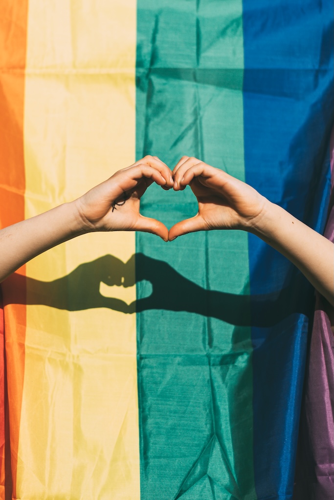 Two hands making a heart in front of a pride rainbow flag