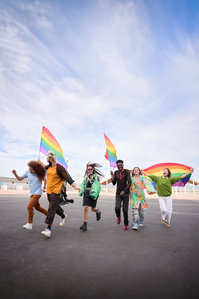 Group happy LGBT people walking down street with rainbow flags under clear sky