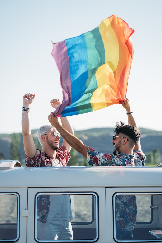 Two young men hugging and holding waving LGBT flag over heads while standing inside retro van with opened roof in nature