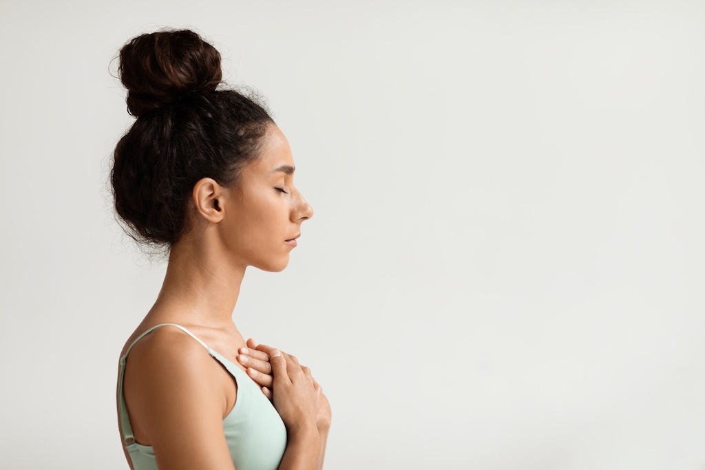Profile portrait of calm young woman with hands on chest and closed eyes, side view shot of peaceful millennial female meditating or praying while standing over white background indoors