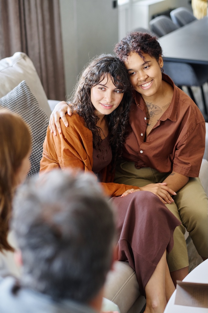 Happy young lesbian couple in casualwear sitting next to each other on couch in front of therapist of one of them during conversation