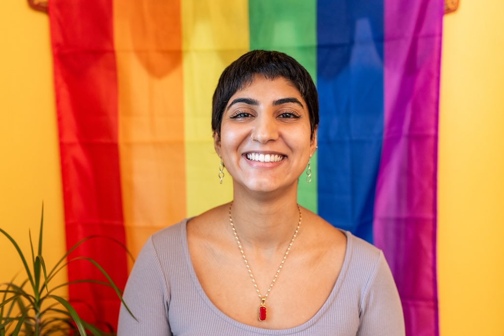 Woman is smiling as she poses for a portrait in front of a rainbow flag.
