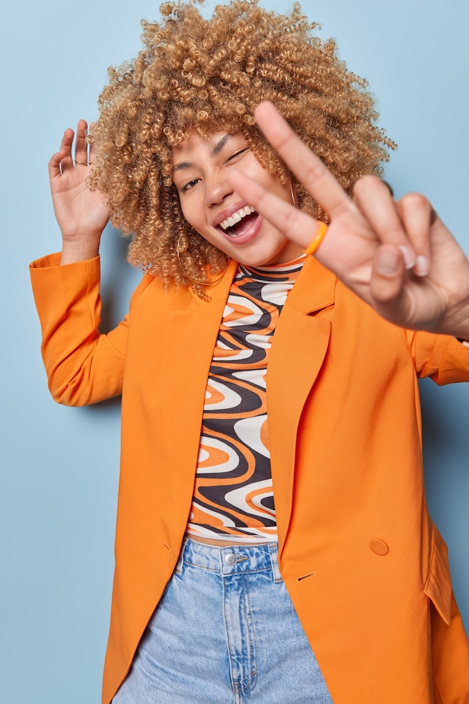 Vertical shot of positive curly haired young woman wears stylish orange jacket and jeans shows peace sign and laughs