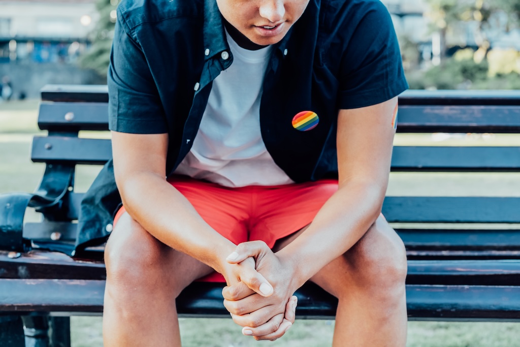 Cropped no face sad LGBTQ indivdual with rainbow badge on t-shirt sitting on park bench alone.