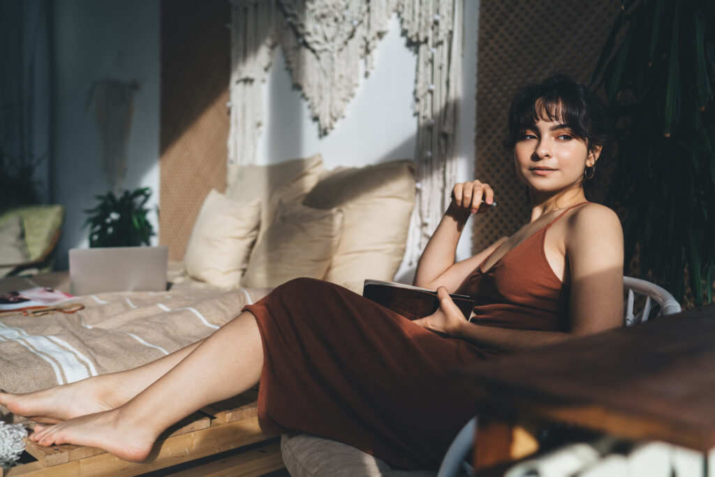 Young calm woman wearing trendy outfit sitting with barefoot on bed while looking away thoughtfully in comfortable bedroom