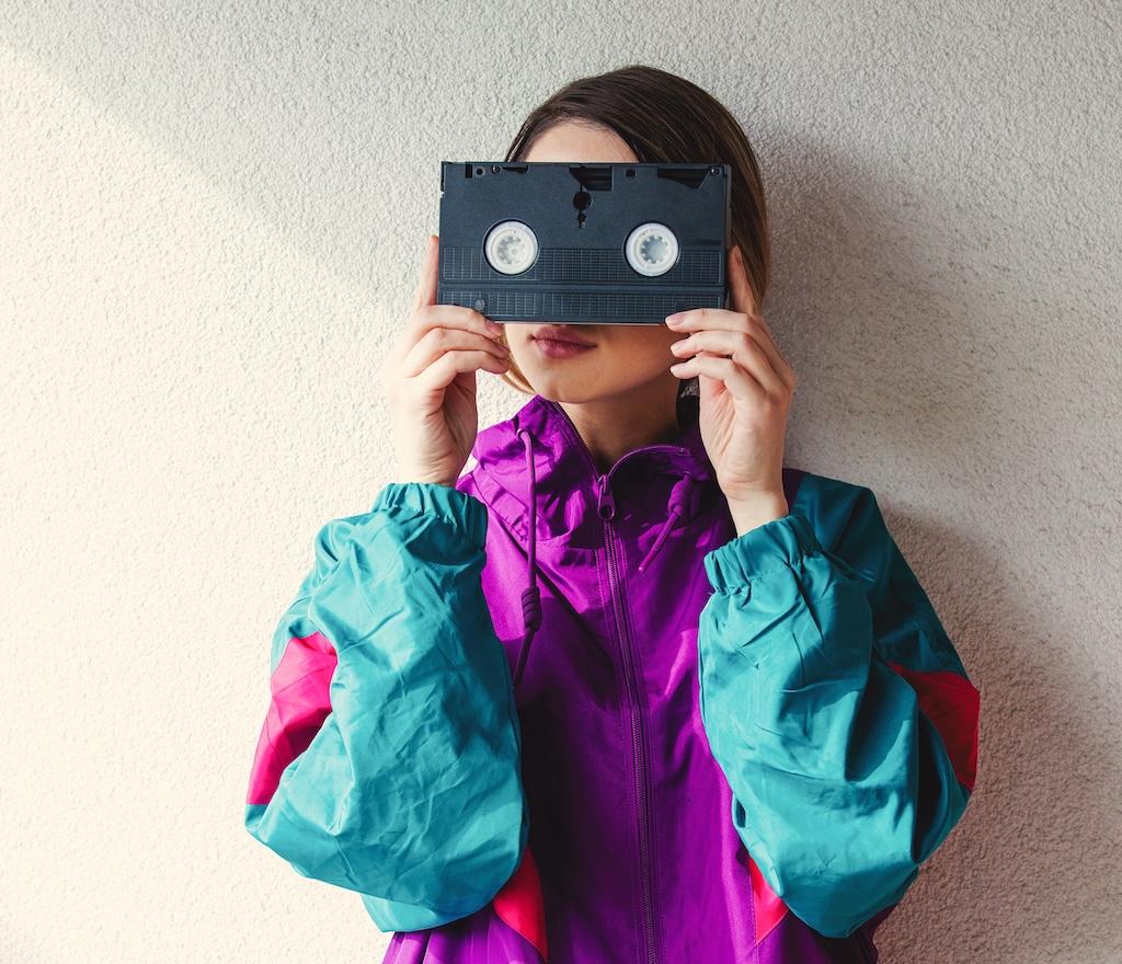 Young woman in 90s style clothes holding VHS cassette on white background
