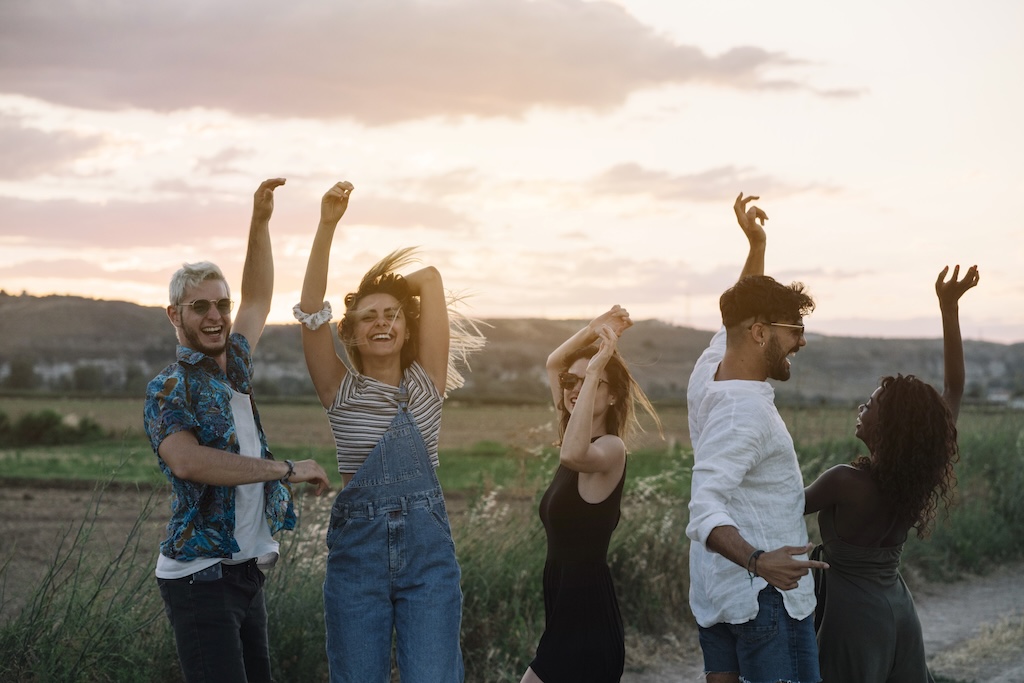 Group of young people in casual outfits laughing and dancing while having fun in beautiful countryside together