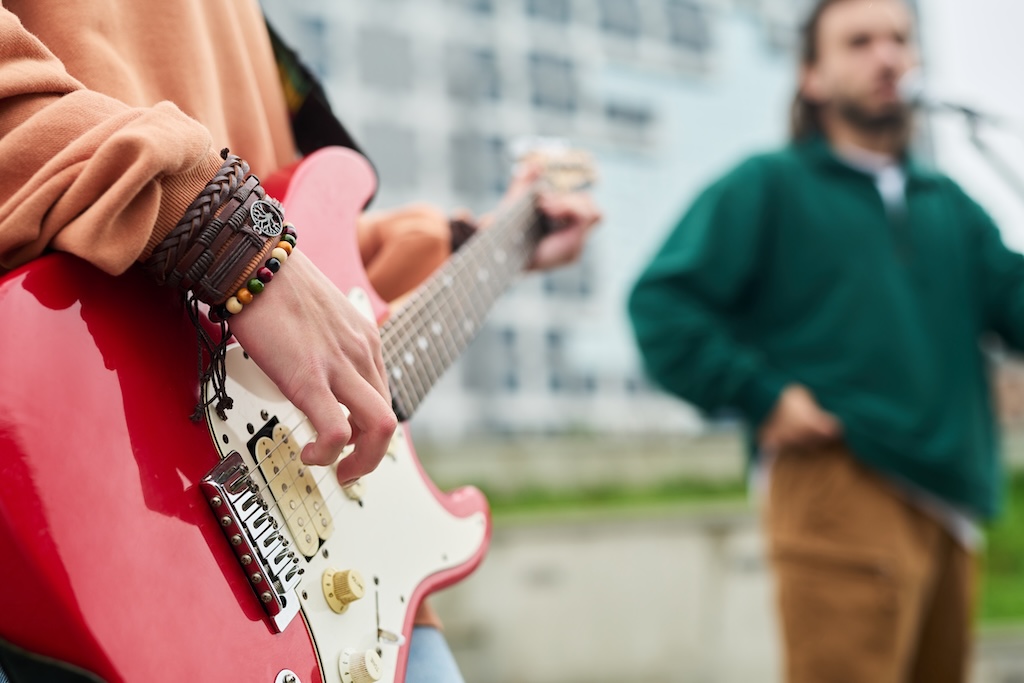 Young adult woman playing electric guitar outdoors with young adult Caucasian man singing in background, focusing on hand strumming strings during live performance