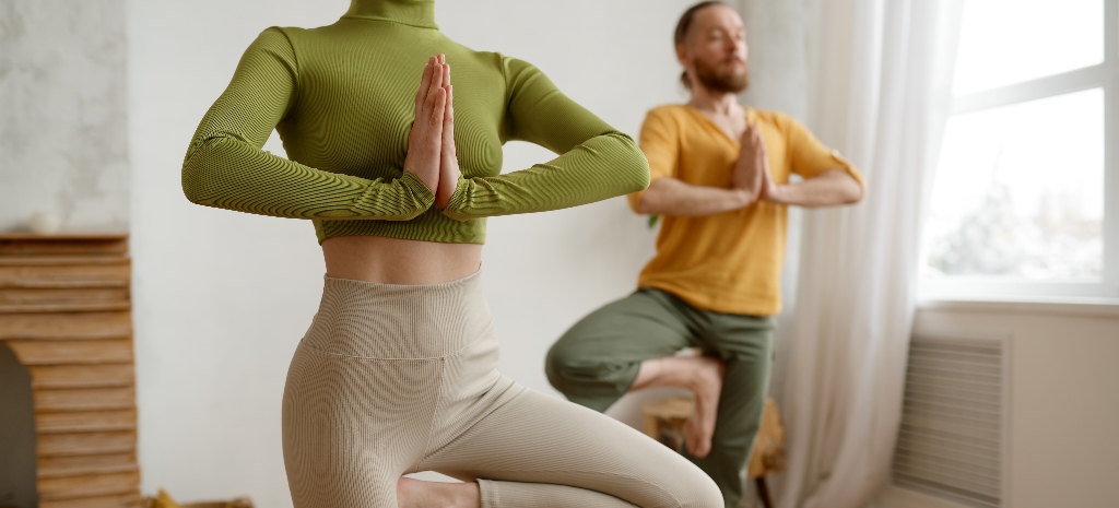 Two people standing in yoga vrikshasana tree pose. Young man and woman wearing sportswear doing balancing exercise. Sport at home. Cropped shot with selective focus 