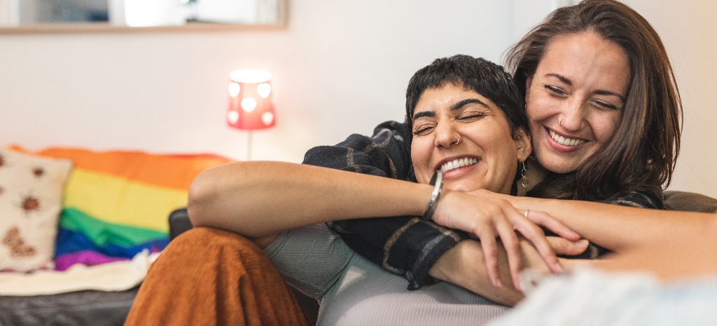 Authentic shot of happy married homosexual female gay couple laughing and embracing on the sofa with rainbow pride flag on background - lesbian couple at home enjoying life together