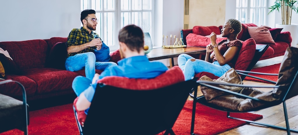 Diverse male and female individuals discussing live socialising during guests meeting in modern living room, youthful hipster guys resting in cosy rehab interior with loft decoration and red furniture 