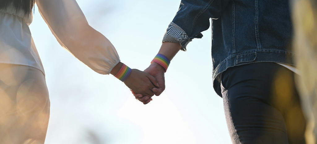 Two lgbtqia community members with rainbow pride flag bracelets holding hands and walking in the sun