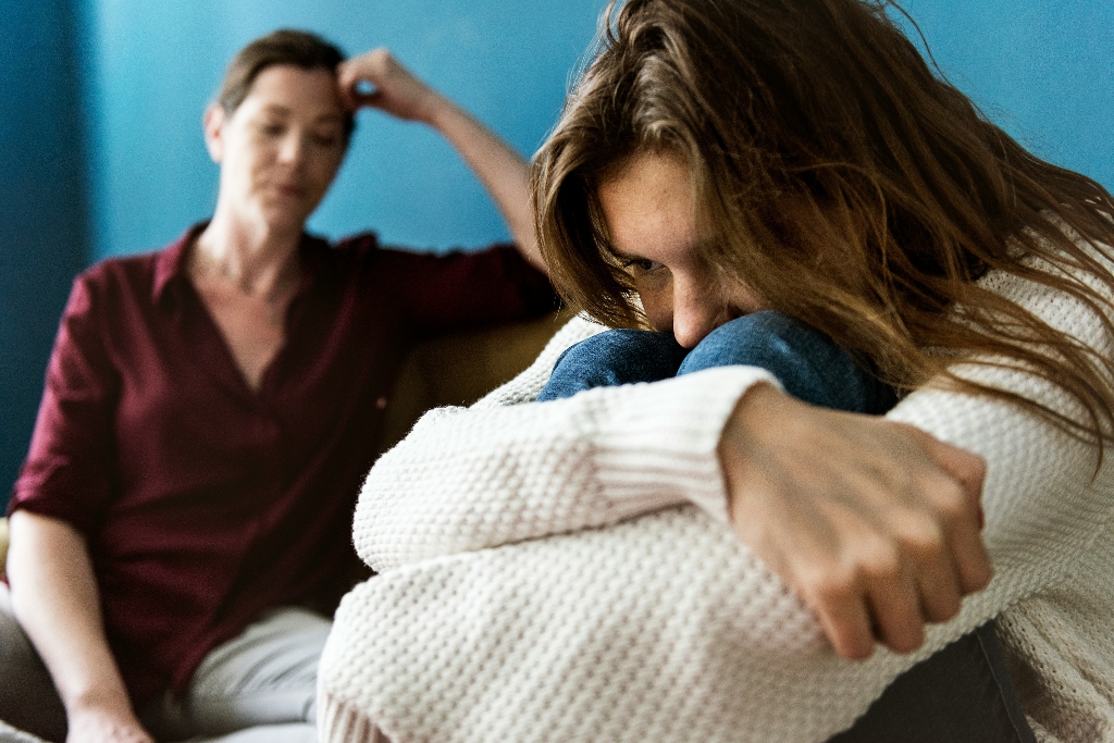 Person sitting curled up looking distressed while another person sits behind them, representing the emotional impact of family rejection and the need for LGBTQIA+-affirming mental health support