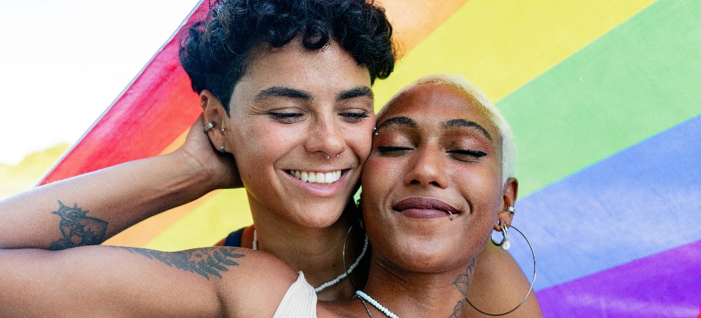 Two people embracing in front of a rainbow flag, smiling and expressing comfort, connection, and support.