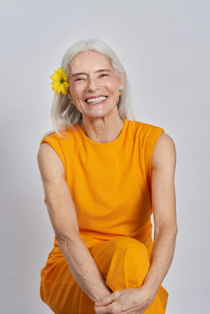 Smiling older woman in an orange outfit with a yellow flower in her hair, representing healing, confidence, and inclusive mental health support.