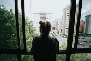 Person standing alone looking out a large apartment window over a quiet cityscape, reflecting themes of isolation, loneliness, and depression.