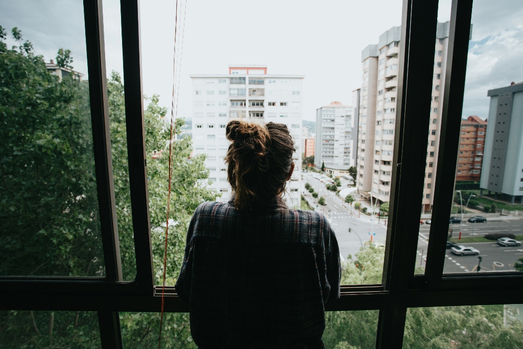 Person standing alone looking out a large apartment window over a quiet cityscape, reflecting themes of isolation, loneliness, and depression.