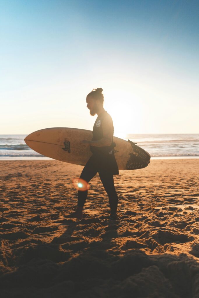Person walking along the beach with a surfboard at sunset, representing the calming and restorative benefits of surf therapy for mental health and trauma recovery.