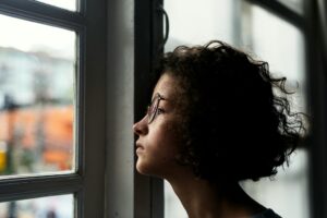 Young adult wearing glasses looking out a window in quiet reflection, representing self-discovery, autism, and gender identity exploration.