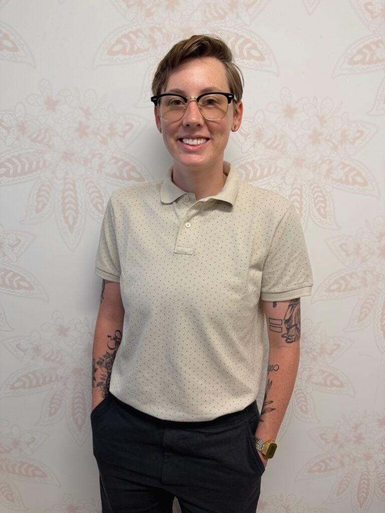 Linzy Holmes, Admissions Coordinator at Element Q Healing Center, standing and smiling in front of a patterned wall wearing glasses, a light-colored collared shirt, and dark pants.