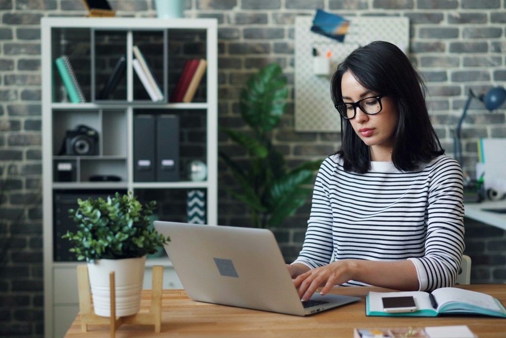Woman wearing headphones and glasses working on a laptop at a desk with a notebook.